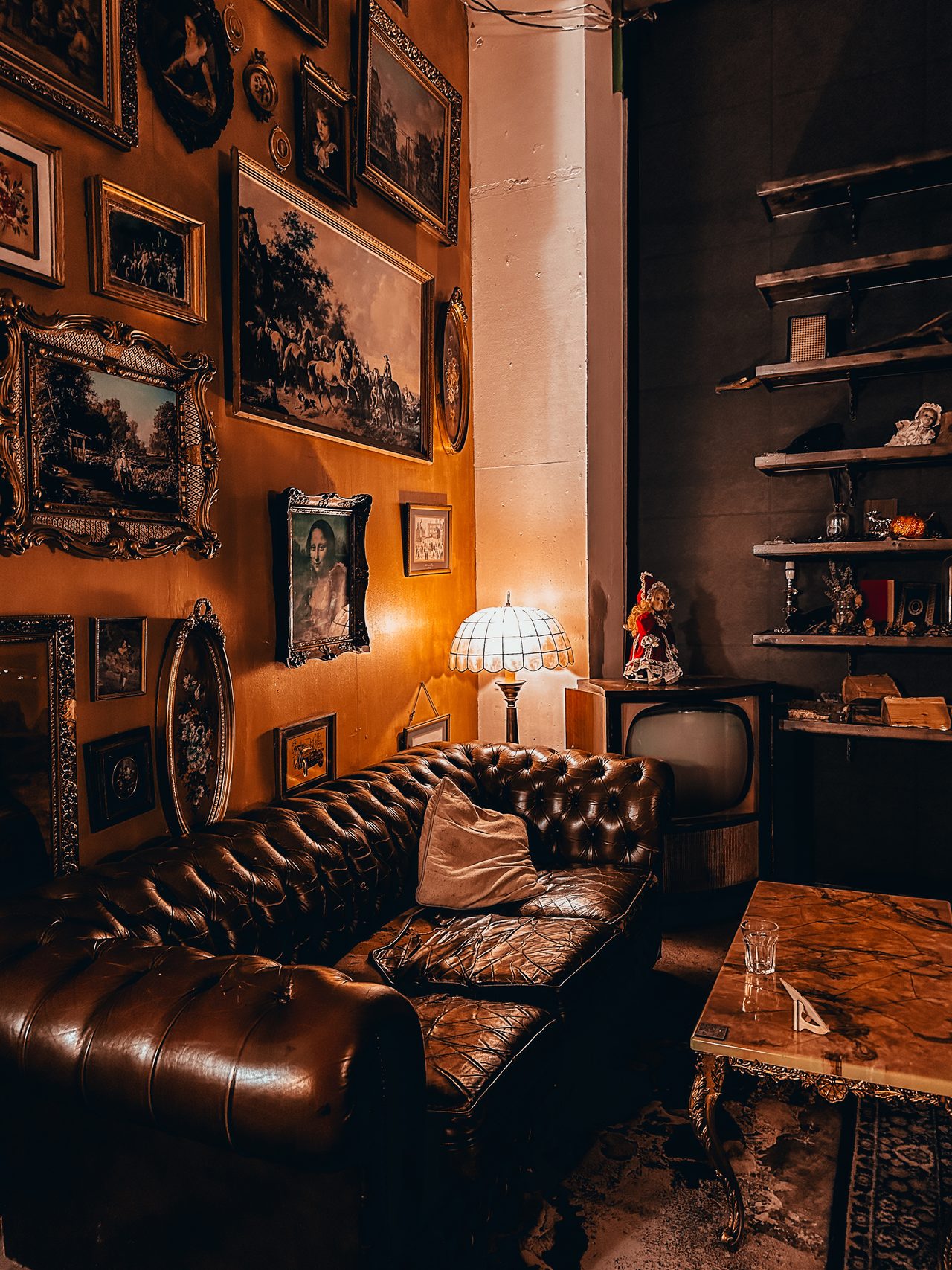 A lamplit lounge of gilt-framed paintings and a Chesterfield sofa